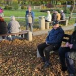 Skupina starejših oseb sedi na klopeh v parku, obdana z odpadlim jesenskim listjem; v ozadju so še drugi stanovalci in osebje, ki se družijo ob sprehajalnih poteh doma za starejše.