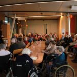 Večja skupina starejših oseb je zbrana ob dolgih mizah, kjer poteka predavanje policista o varnosti v vsakdanjem življenju.