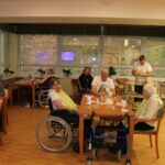 Stanovalci med zajtrkom v jedilnici doma, nekateri sedi za mizami, negovalka v ozadju pripravlja hrano.