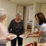 Dve stanovalki prejemata kos kruha z maslom od zaposlene, ki jima ga razreže in postreže.