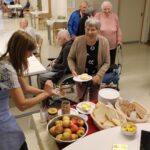 Stanovalci stojijo in sedijo ob postrežni mizi, zaposleni mažejo med na kruh, v ospredju skleda jabolk in narezani kruh.