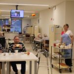 Stanovalci jedo tradicionalni zajtrk za mizami na oddelku, v ospredju negovalka ob postrežnem vozičku z živili in posodami.