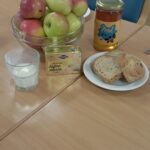 Skleda rdeče-zelenih jabolk, kozarec medu, zavitek masla, kozarec mleka in krožnik z rezinami kruha, razporejeni na leseni mizi v jedilnici doma za starejše.