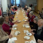 Skupina starejših oseb sedi ob dolgi jedilni mizi, na kateri so skodelice, krožniki s kruhom, maslom in medom; stanovalci se pogovarjajo in zajtrkujejo.