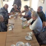 Starejši moški in ženske sedijo okoli manjše pravokotne mize, jedo kruh z maslom in medom ter se družijo; ob mizi so skodelice, krožniki in pripomočki za zajtrk.