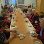 Številna skupina starejših oseb sedi ob zelo dolgi jedilni mizi z jabolki, kruhom, maslom in toplim napitkom; udeleženci se pogovarjajo in uživajo tradicionalni slovenski zajtrk v skupnem prostoru.