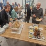 Skupina starejših oseb si pri mizi pripravlja prigrizke iz kruha, masla in medu.