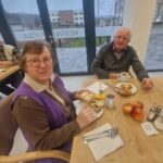 Par starejših oseb sedi za mizo in uživa tradicionalni zajtrk s kruhom, maslom in medom.