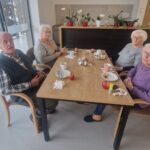 Majhna skupina starejših oseb sedi za mizo in se pogovarja ob jutranjem napitku in sadju.