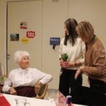 Stanovalka, ki praznuje 100 let, prejema rožo in voščila od zaposlenih in obiskovalcev; za njo je okrašena stena s številko 100.