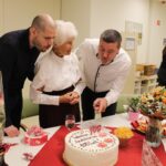 Jubilantka z dvema moškima gleda praznično torto z napisom za 100. rojstni dan; miza je pogrnjena s prigrizki in dekoracijo.