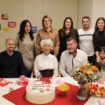 Slavljenka za 100. rojstni dan sedi ob torti, obdana z družinskimi člani, ki se skupaj fotografirajo; miza je praznično obložena s cvetjem, prigrizki in pijačo.