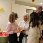 Skupina svojcev in zaposlenih čestita stanovalki ob njenem 100. rojstnem dnevu; v ozadju so zlati baloni in številka 100, slavljenki pa predajajo voščilnico.