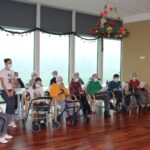 Stanovalci doma sedijo v polkrogu v svetlem prostoru in berejo besedilo, ob njih stoji spremljevalka; prostor je praznično okrašen z božičnimi okraski.