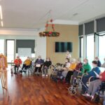 Skupina stanovalcev, večinoma z uporabo pripomočkov za gibanje, sedi in spremlja branje ali predstavitev v skupnem prostoru z velikimi okni in praznično dekoracijo.