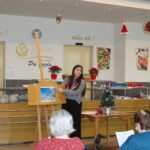 Spremljevalka stoji ob stojalu s slikanico in prebira ali pripoveduje zgodbo stanovalcem, ki sedijo pred njo v jedilnici doma.