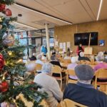 Starejši sedijo v jedilnici doma za starejše in poslušajo branje ob božični dekoraciji.