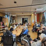 Več starejših stanovalcev, tudi na invalidskih vozičkih, sedi za mizami v skupnem prostoru doma za starejše.