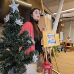 Oseba bere besedilo ob lesenem stojalu, okrašenem z božično dekoracijo, v skupnem prostoru doma za starejše.