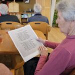 Starejša ženska sedi za mizo in bere natisnjeno besedilo med skupinskim dogodkom v domu za starejše.