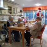 Stanovalci doma za starejše sedijo za mizo v skupni kuhinji, Božiček pa jih obišče in jim nameni pozdrav.