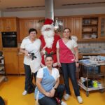 Božiček stoji skupaj z zaposlenimi doma za starejše v kuhinji in se fotografira ob vozičku z opremo.