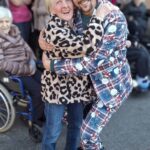 Stanovalka doma za starejše in obiskovalec se nasmejana objemata pred vhodom v stavbo.