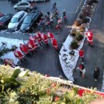 Skupina obiskovalcev, oblečenih v Božičke, stoji na dvorišču pred domom; ob njih so parkirana kolesa in motorji.