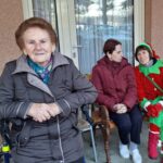 Stanovalka doma za starejše sedi na klopi pred stavbo, v ozadju sedita še dve osebi, ena v božičnem kostumu.