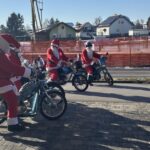 Skupina motoristov, oblečenih v Božičke, se vozi z motorji po cesti na sončen zimski dan.