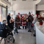 Skupina starejših ljudi v domu za starejše, ki jih obiščejo Božički in škrat z glasbo; nekateri stanovalci so na invalidskih vozičkih.