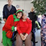 Božični škrat objema starejšo žensko na invalidskem vozičku v skupnem prostoru doma za starejše.