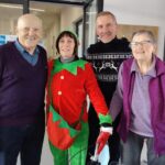 Štirje nasmejani starejši in božični škrat pozirajo v notranjem prostoru doma za starejše.