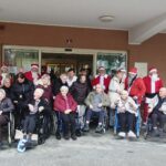 Skupinska fotografija stanovalcev doma za starejše na invalidskih vozičkih in spremljevalcev, pred vhodom v stavbo; ob njih stojijo obiskovalci, oblečeni v Božičke.