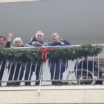 Stanovalci doma za starejše stojijo in mahajo z balkona, okrašenega z božičnim okrasjem iz zelenja.