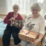 Dve starejši ženski sedita na postelji in držita zaviti božični darili v sobi doma za starejše.