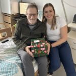 Starejši moški in mlajša ženska sedita na postelji ter skupaj držita zavito božično darilo.