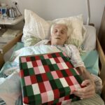 Starejša oseba leži v postelji in drži veliko zavito božično darilo.