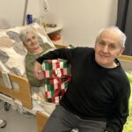 Starejši par v sobi doma za starejše drži zavito božično darilo in gleda proti kameri.