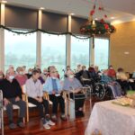 Stanovalci, večinoma sedeči na stolih in invalidskih vozičkih, zbrani pri božični sveti maši v svetlem prostoru z velikimi okni.