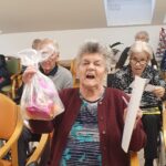 Stanovalka sedi na stolu, v roki drži vrečko z nagrado in listek ter veselo sodeluje v božični tomboli.