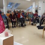 Zaposlena sedi na stolu pred skupino stanovalcev in vodi božično tombolo v svetlem prostoru doma.