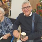 Starejša gospa in moški sedita drug ob drugem na prireditvi, pijeta topel napitek in se nasmihata. Za njima stoji božično drevo.