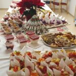 Miza, bogato obložena s prigrizki, pecivom, narezki in sladkimi kozarčki. Na sredini stojita dve božični zvezdi in dekorativno zloženi prtički.