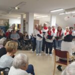 Skupina pevk v belem oblačilu z rdečimi šali poje ob električnem klavirju. Okoli njih sedi skupina stanovalcev doma, ki spremljajo nastop.