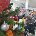 Stanovalci doma sedijo v polkrogu in spremljajo dogodek. V ospredju je okrašena božična smrečica z rdečimi in srebrnimi okraski.