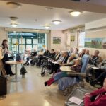 Predavateljica stoji ob slikarskem stojalu in bere v mikrofon, stanovalci doma za starejše pa jo sede poslušajo v dnevnem prostoru.