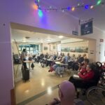 Pogled iz hodnika v dnevni prostor, kjer predavateljica bere, pred njo pa sedi večja skupina stanovalcev doma za starejše.