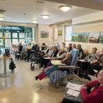 Stanovalci doma za starejše sedijo v vrstah in poslušajo branje, predavateljica stoji ob slikarskem stojalu.