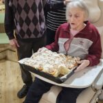 Starejša gospa sedi na stolu in drži pekač s pečenimi cimetovimi rolicami, ob njej stojita starejši gospod in gospa ter jo opazujeta.